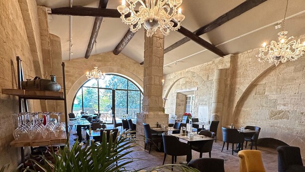 A spacious dining area with high ceilings, exposed beams, and large windows. Tables are set with chairs, and a chandelier hangs from the ceiling. - Domaine du Prieuré d'Estagel (Saint-Gilles)