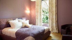 A bedroom with a bed, a chair, a window with curtains, and a wall-mounted light. - Domaine du Prieuré d'Estagel (Saint-Gilles)