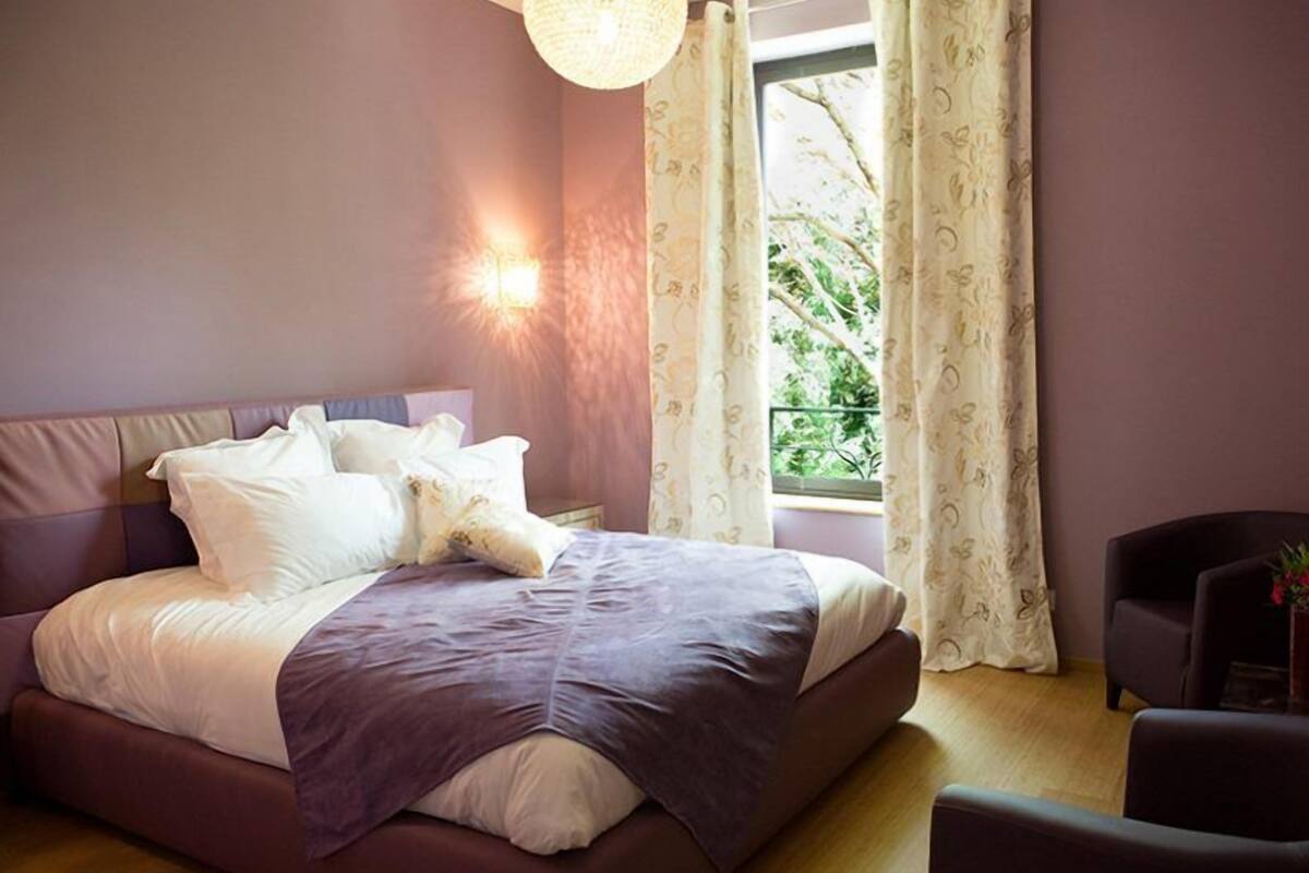 Een slaapkamer met een bed, een stoel, een raam met gordijnen en een wandlamp.