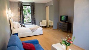 A modern bedroom with a bed, a blue sofa, a television, and a small table with a vase of flowers. - Domaine du Prieuré d'Estagel (Saint-Gilles)
