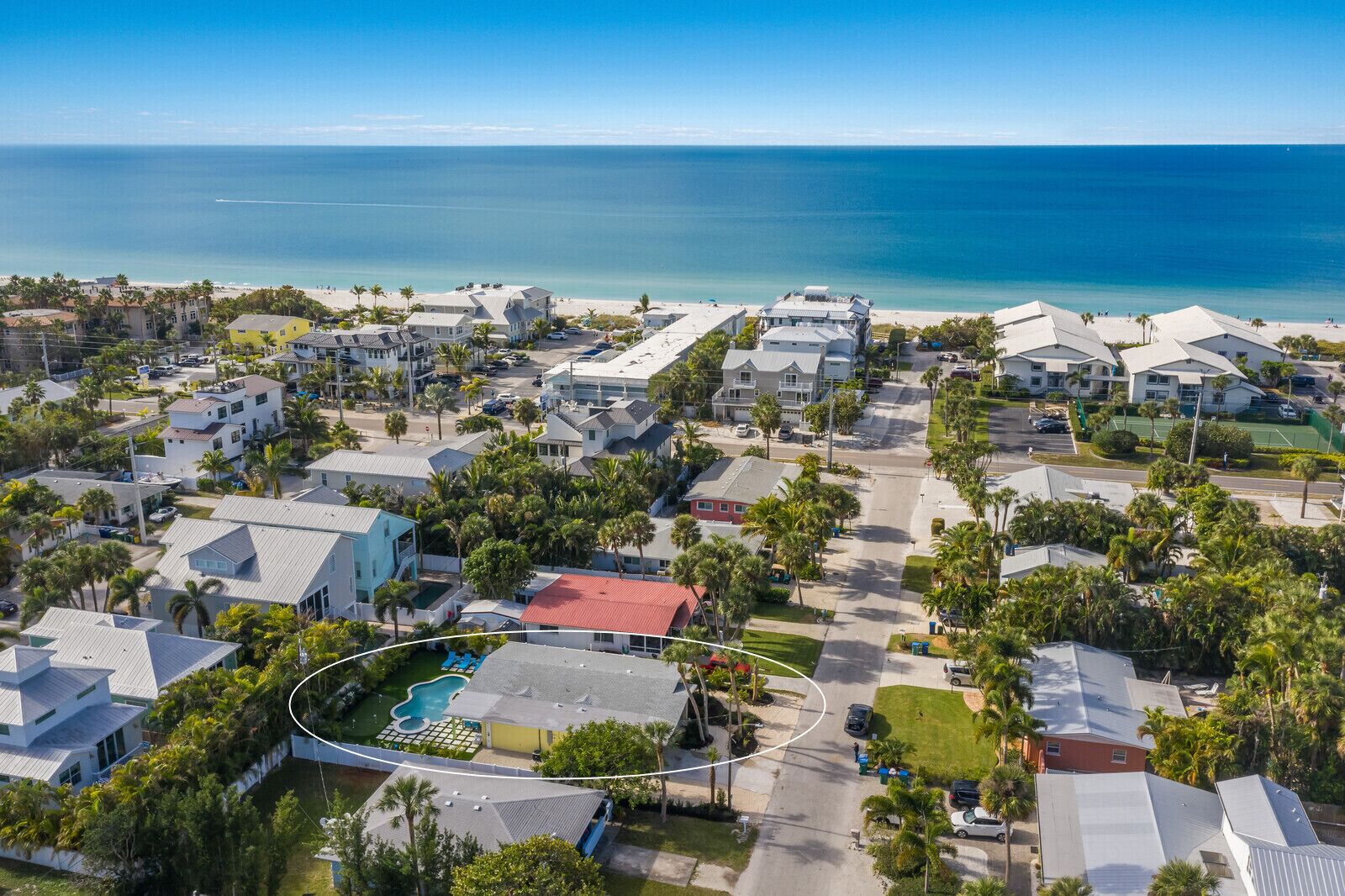 An aerial view of a coastal residential area with houses, a swimming pool, and a beach in the background.