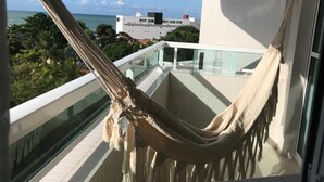 Un balcón con hamaca, barandillas de vidrio y vista al océano y edificios.