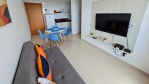 Una sala de estar moderna con un sofá gris, una mesa de comedor con sillas azules, una cocina al fondo y un televisor montado en la pared.