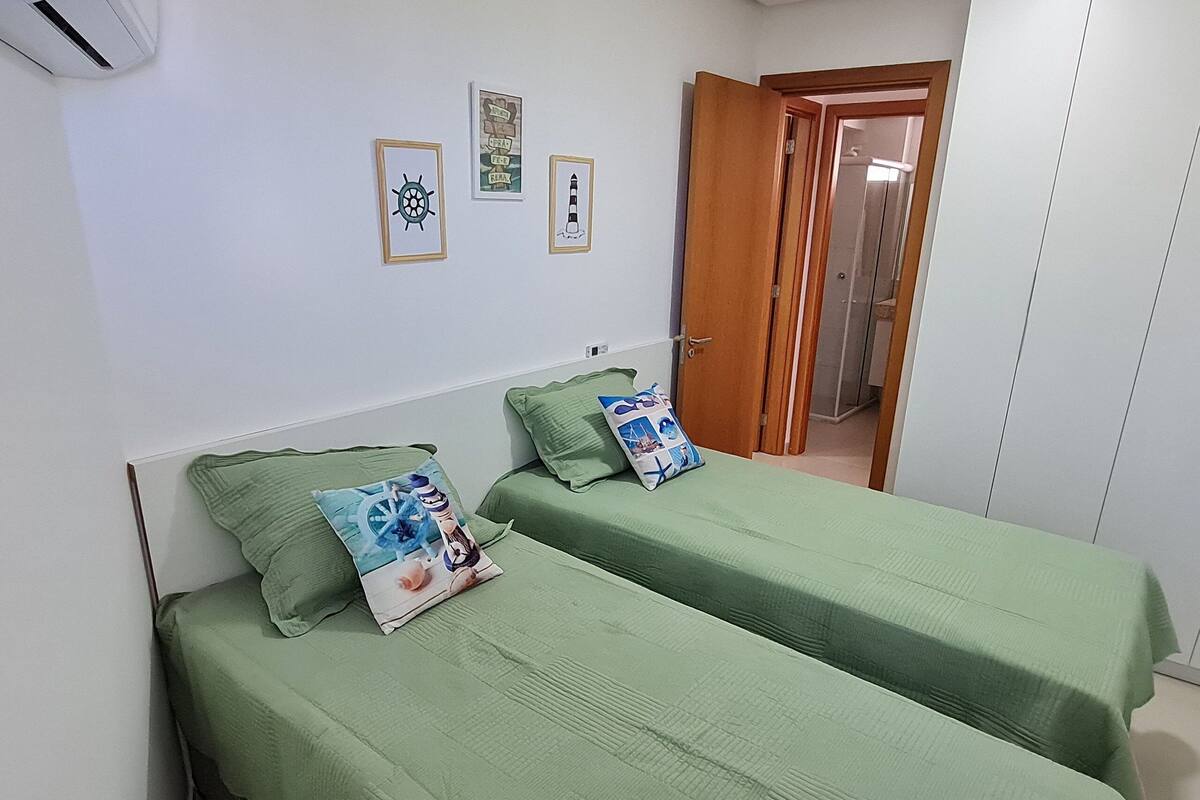 A room with two single beds, each with a green bedspread and matching pillows. There are framed pictures on the wall above the beds.