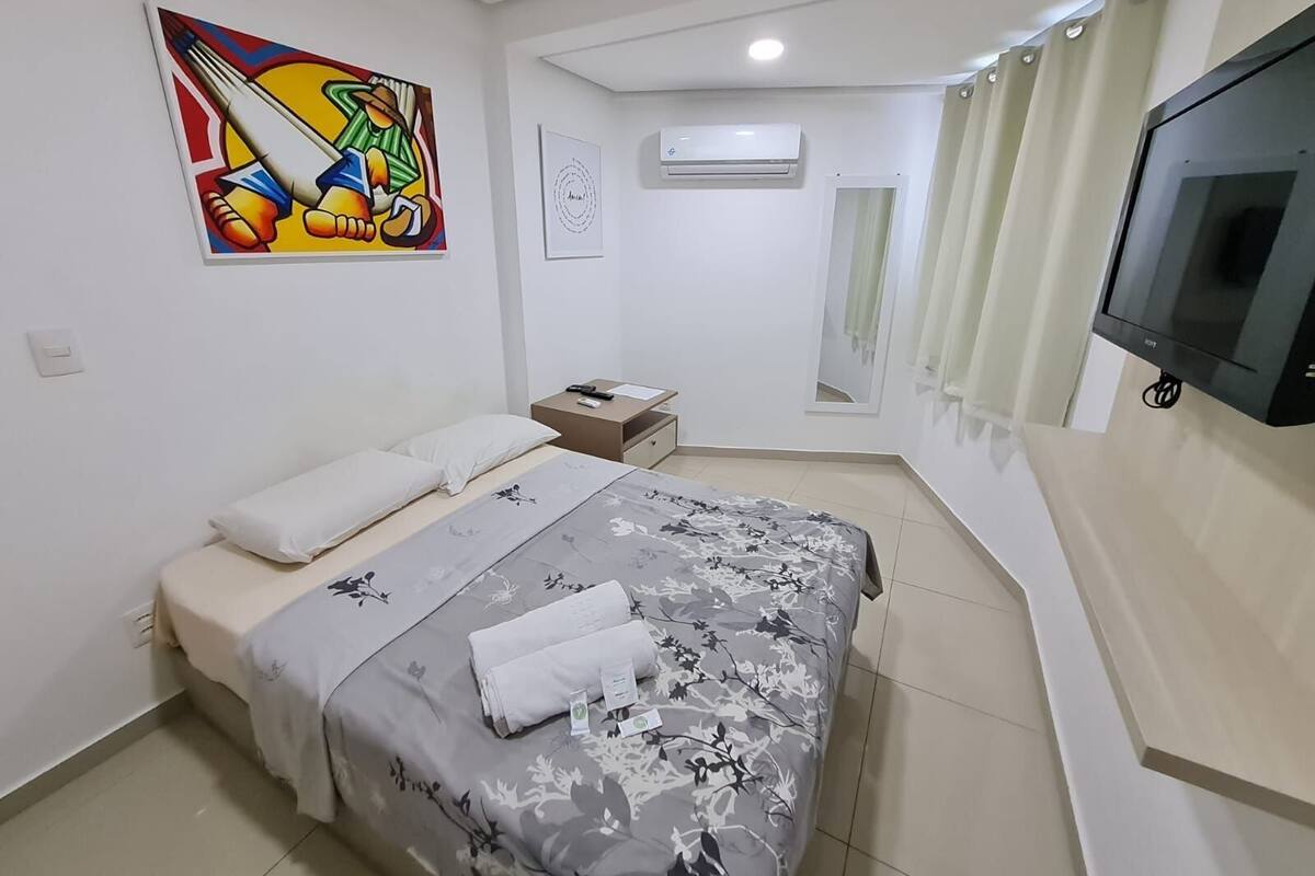 A bedroom with a bed, a nightstand, an air conditioning unit, a TV, and a painting on the wall.