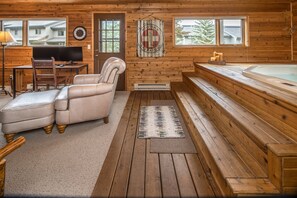 Interior de una cabaña de madera con piso de madera, un sofá de cuero, un escritorio con una computadora, un televisor, una chimenea y un jacuzzi.