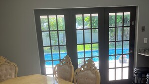 A dining area with a large table and chairs, a view of a swimming pool through a glass door, and a tiled floor. - Relaxing 3 bedroom pool home (Miami)
