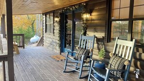 Une terrasse en bois avec deux fauteuils à bascule et une petite table, une cheminée en pierre et une vue sur les arbres environnants.