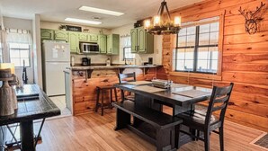 Une cuisine de style rustique, aménagée comme une cabane, avec des murs en bois, une table à manger avec des chaises, un réfrigérateur, un four à micro-ondes et un lustre.