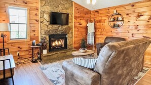 L’intérieur d’un chalet confortable, avec des murs en rondins, une cheminée en pierre, un téléviseur à écran plat fixé au-dessus, un canapé confortable et une petite table sur laquelle est posé un bol rempli de pommes de pin.