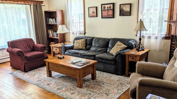 A living room with a leather sofa, a wooden coffee table, a red armchair, a bookshelf, and a patterned rug. - Charming 2BR Private Home in Downtown Bellefonte • Near Penn State & I-80 (Bellefonte)