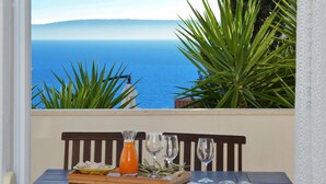Una mesa con una botella de jugo de naranja, dos vasos y un plato de comida, con vista a la costa, plantas verdes y un banco.