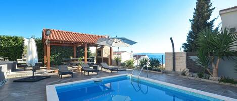 A swimming pool with lounge chairs and umbrellas, a pergola, and a view of the sea.