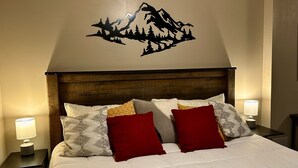 Een net opgemaakt bed met een muurdecoratie met bergthema, een houten hoofdbord en twee bedlampjes.