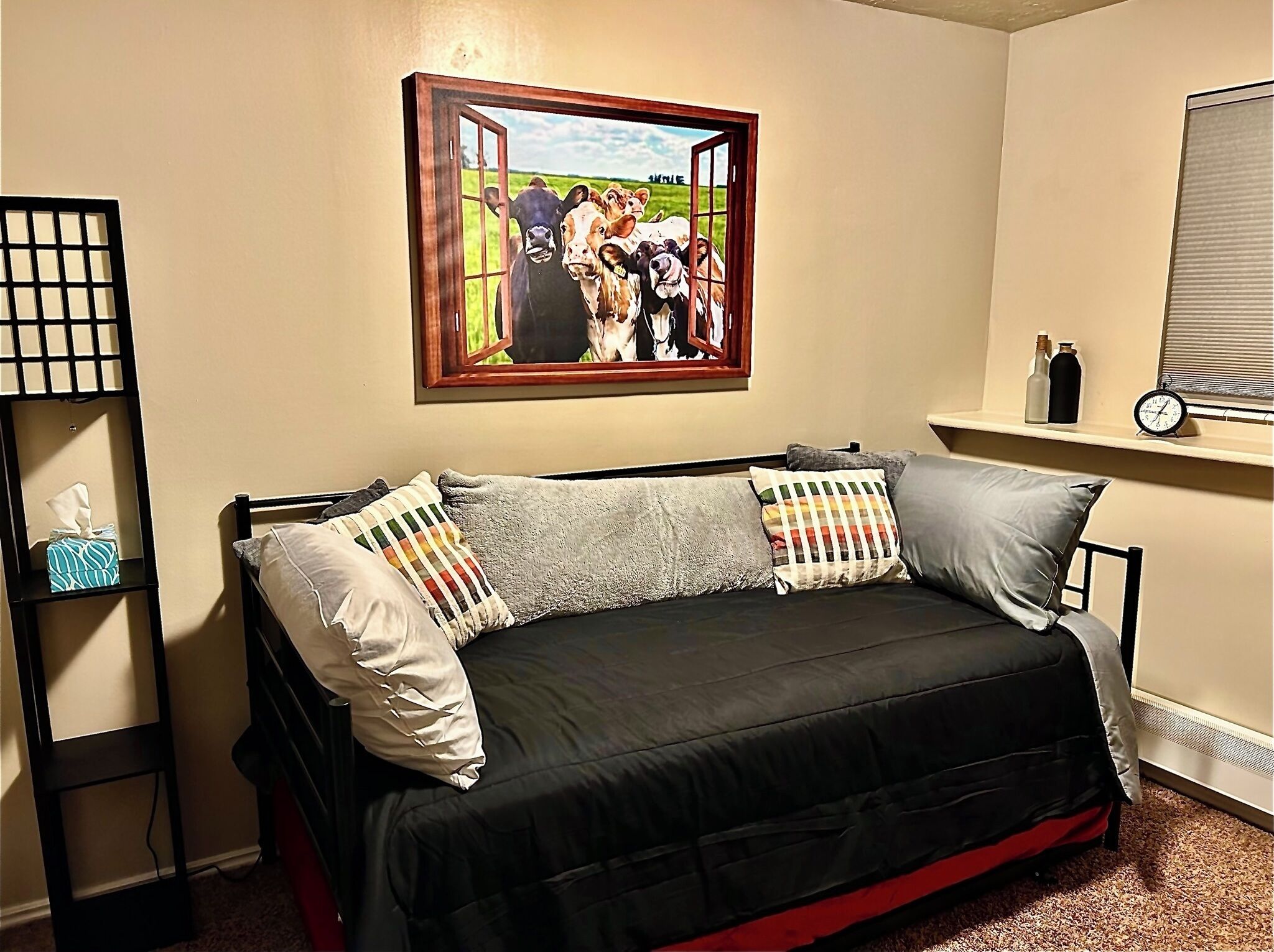 Una habitación con un sofá cama negro, un cuadro de vacas en la pared, una estantería con botellas y un reloj.