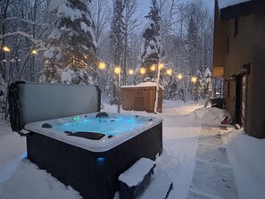A snow-covered outdoor area with a hot tub, wooden cabin, and string lights.