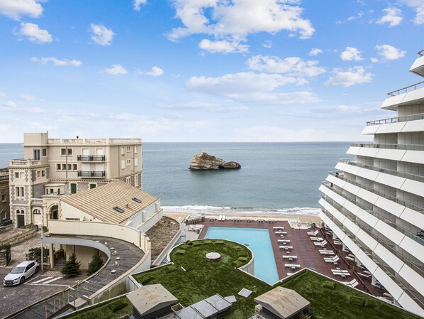 A modern hotel with a rooftop pool, a view of the sea, and a rocky outcrop in the distance.