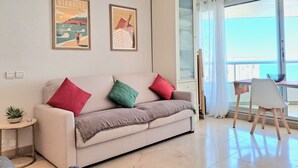 A living room with a sofa, two cushions, a small table with a plant, and a window with a view of the sea. - Studio Roche Ronde - Welkeys (Biarritz)