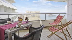 A balcony with wicker furniture, a table set with a bowl of fruit, and a view of the sea and buildings. - Studio Roche Ronde - Welkeys (Biarritz)