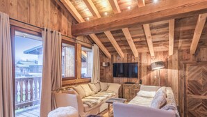 Hyggelig interiør i en træhytte med sofa, to skottomaner, fjernsyn og en balkon med gardiner.