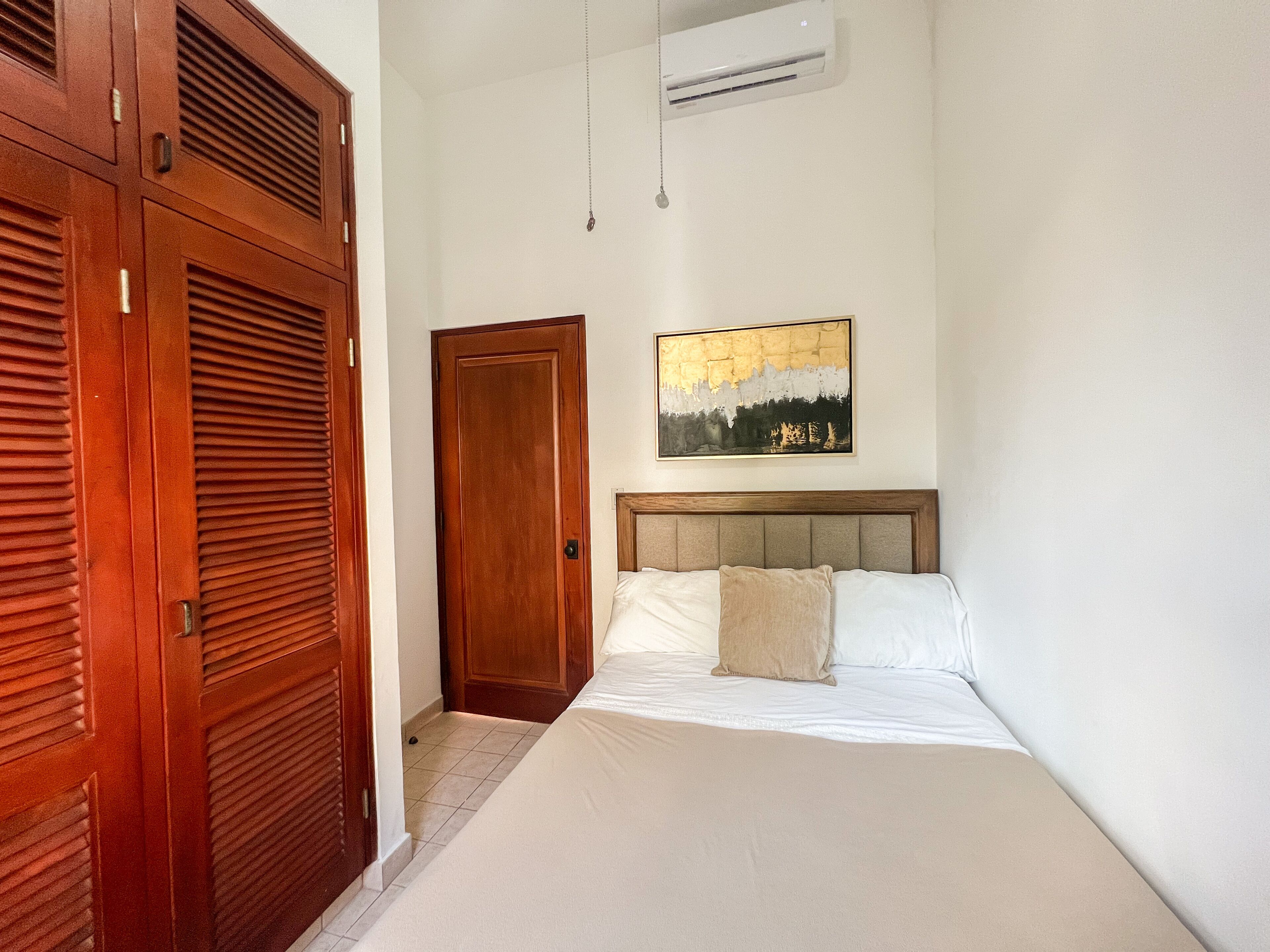 A bedroom with a bed, a wooden wardrobe, a door, and a wall-mounted air conditioner.