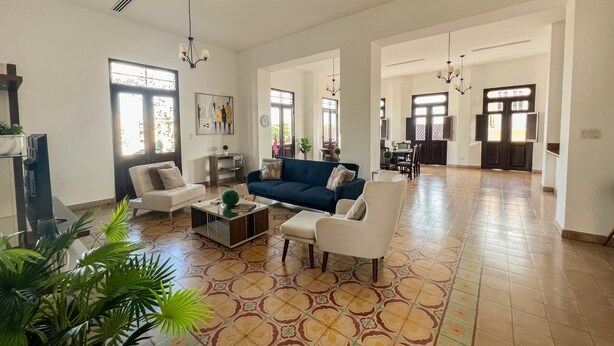 Amplia sala de estar con un sofá azul, sillones blancos y una mesa de centro. Hay un televisor a la izquierda y un comedor al fondo.