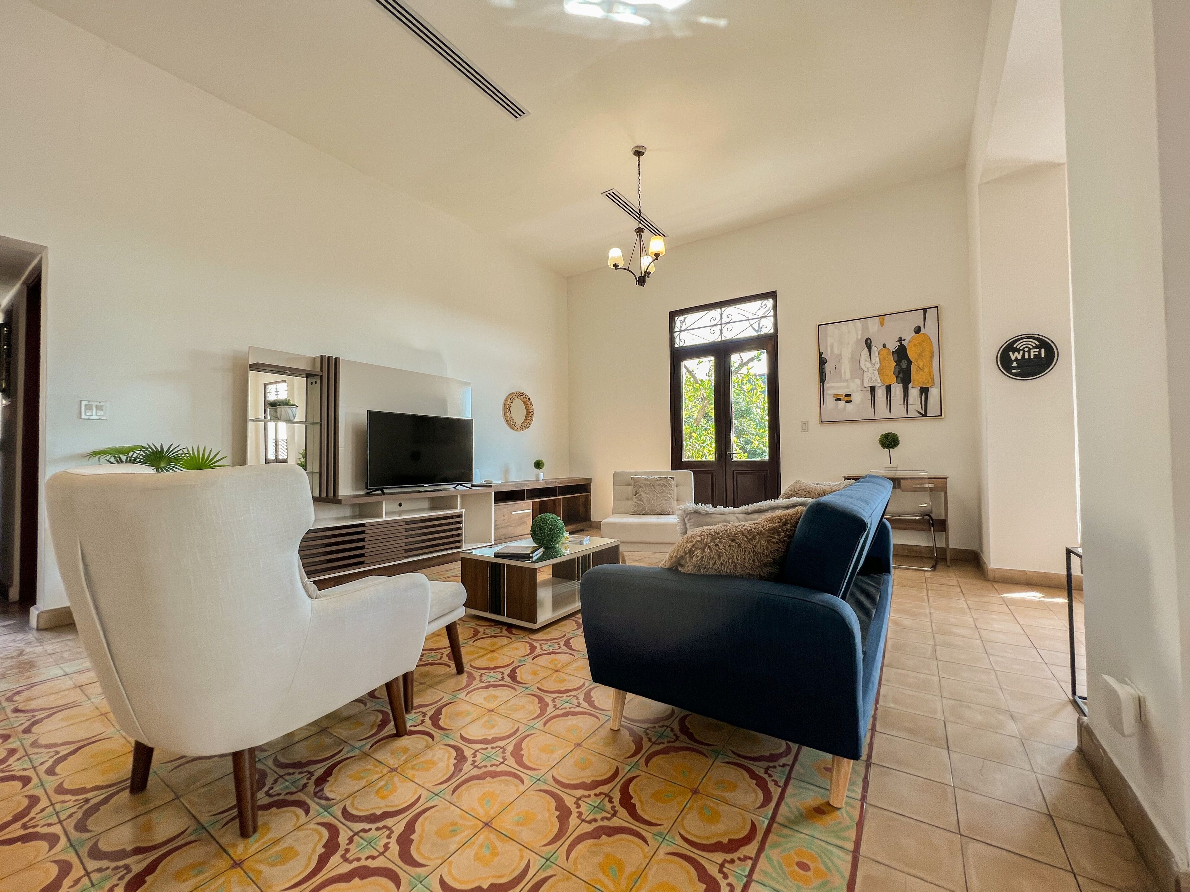 Amplia sala de estar con televisor grande, sofá, sillón y mesa de centro. Se ve una ventana con vista a árboles y una puerta con marco de metal decorativo.
