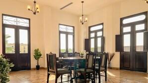Un comedor con una mesa redonda, sillas, plantas en macetas y ventanas grandes con contraventanas.