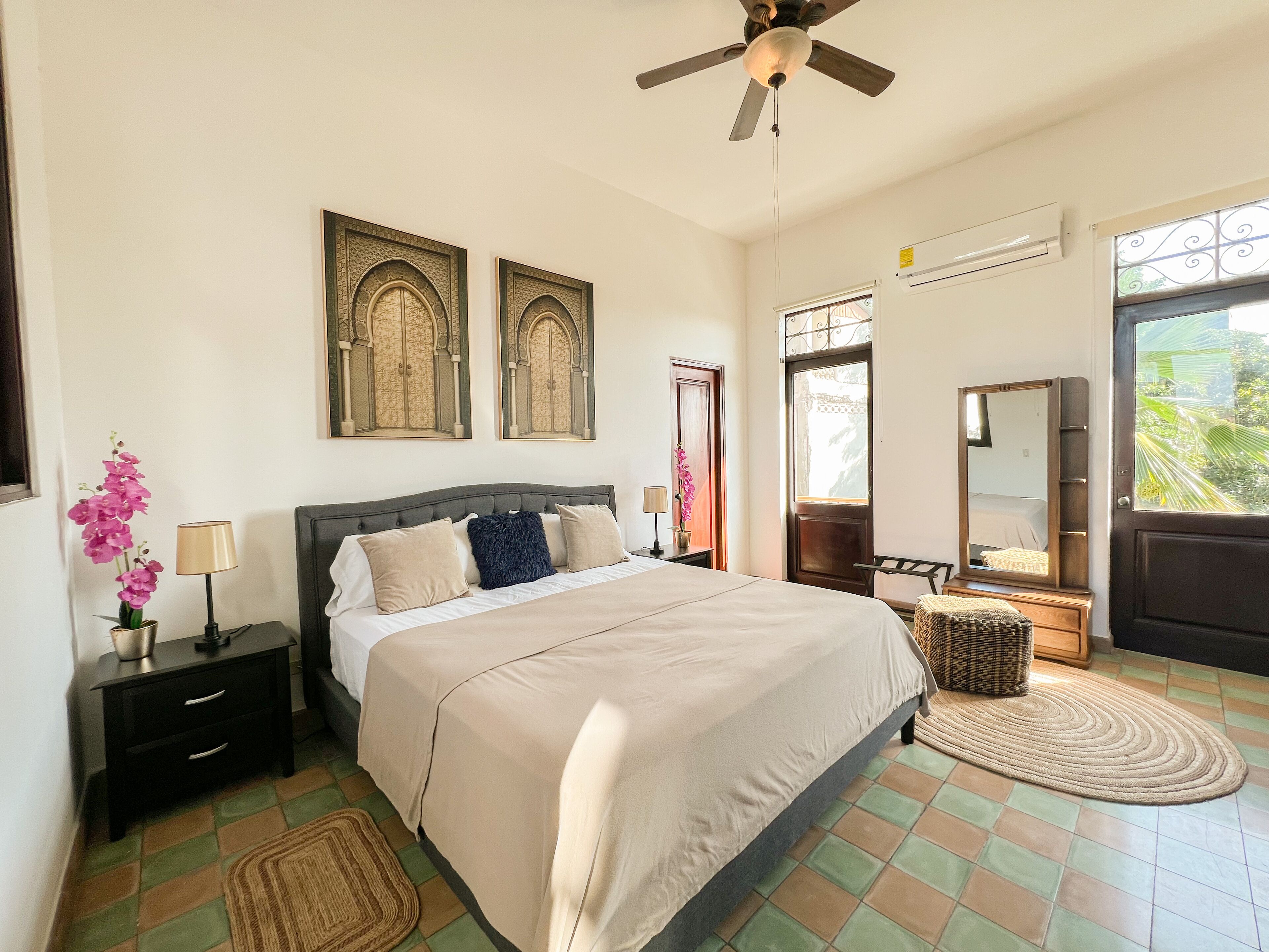 A bedroom with a bed, nightstands, a ceiling fan, a window with a view, and decorative elements.