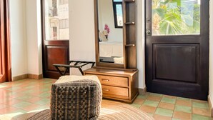 Una habitación con un aparador de madera, un espejo, un reposapiés de mimbre y vistas de un edificio a través de una ventana.