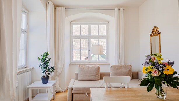 A bright, minimalist living room with a sofa, two armchairs, a small table, a potted plant, a mirror, and a vase of flowers.