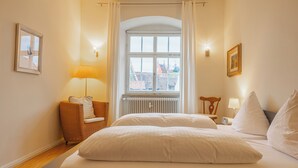 A bedroom with a large bed, a window with curtains, a chair, and a lamp.