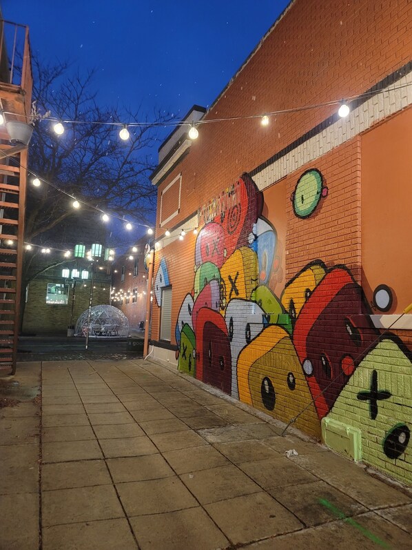 A brick alley with a colorful mural on the wall, string lights, and a metal structure.