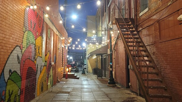 Un callejón estrecho con edificios de ladrillo, un mural en la pared de la izquierda y luces colgantes arriba.