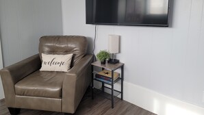 Un sillón de cuero marrón con una almohada que dice 'bienvenido', una mesita con una lámpara y libros, y un televisor de pantalla plana montado en la pared.