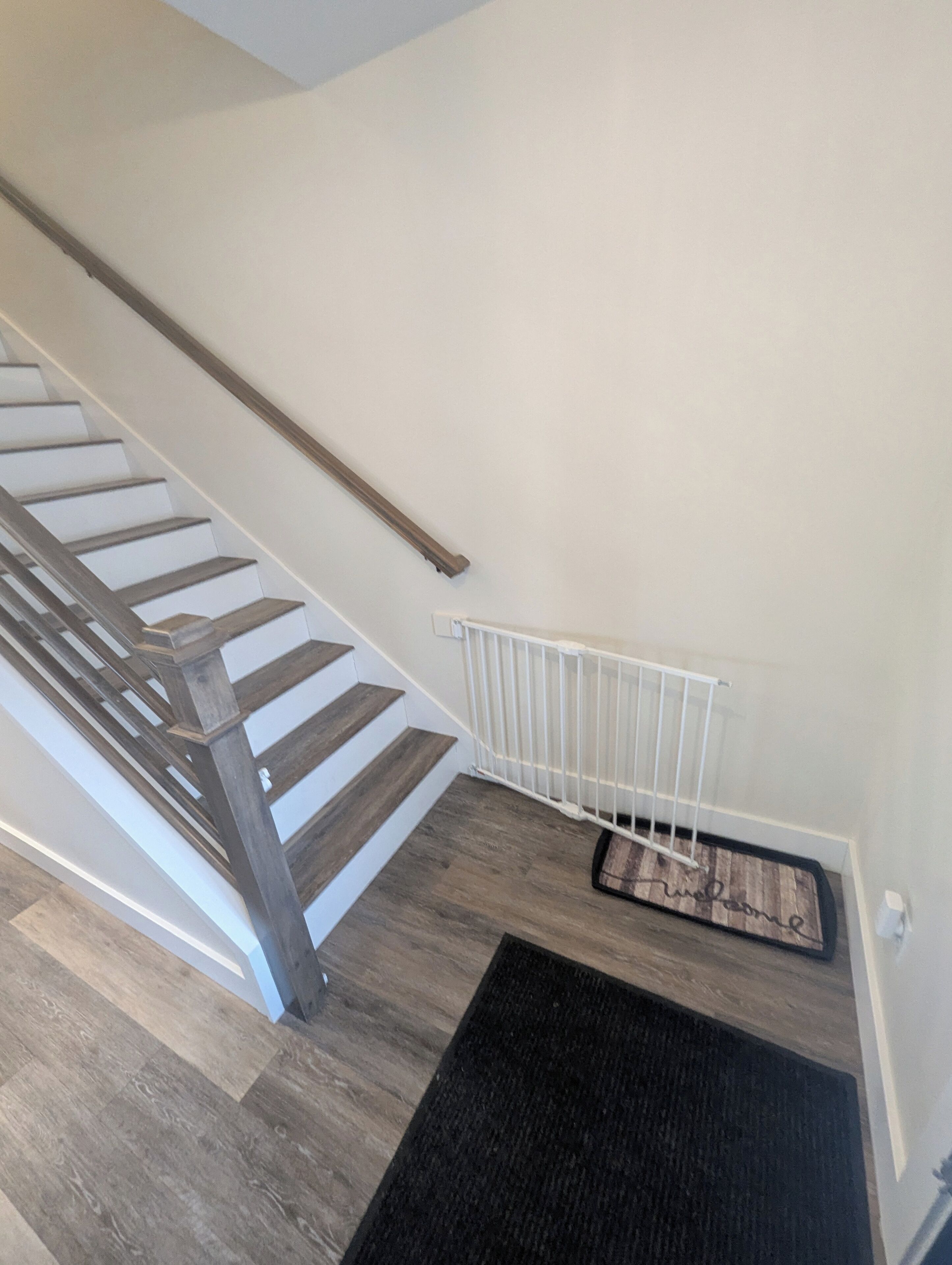 Un escalier avec des marches en bois et une rampe blanche, menant à un palier où se trouvent un paillasson noir et un radiateur blanc.