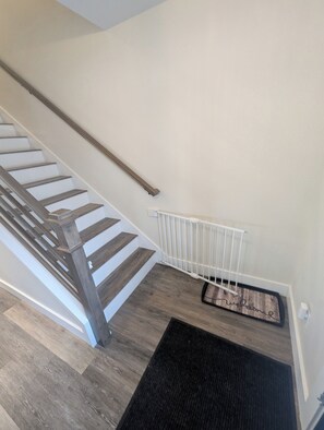 Una escalera con escalones de madera y barandilla blanca, que lleva a un rellano con un felpudo negro y un radiador blanco.