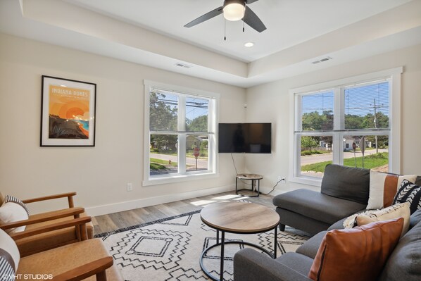 A spacious living room with a large window, a ceiling fan, a flat-screen TV, and a framed picture on the wall.