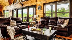 A cabin interior with two leather sofas, a coffee table with food, and large windows offering a view of the outdoors. - Mountain View l 3mi to Gatlinburg l Hot Tub l Game Room l Pack N Play (Gatlinburg)