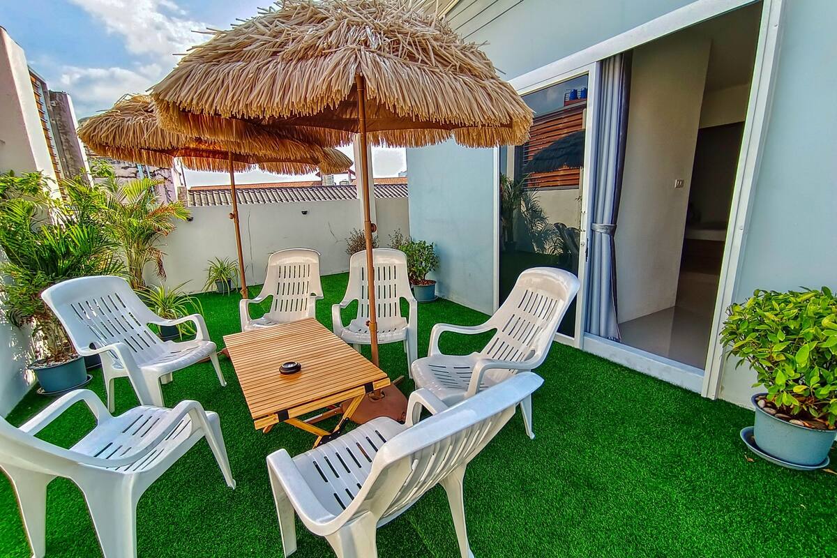 Un espace extérieur aménagé avec des chaises en plastique blanc et une table en bois, entouré de gazon artificiel et de plantes en pot.