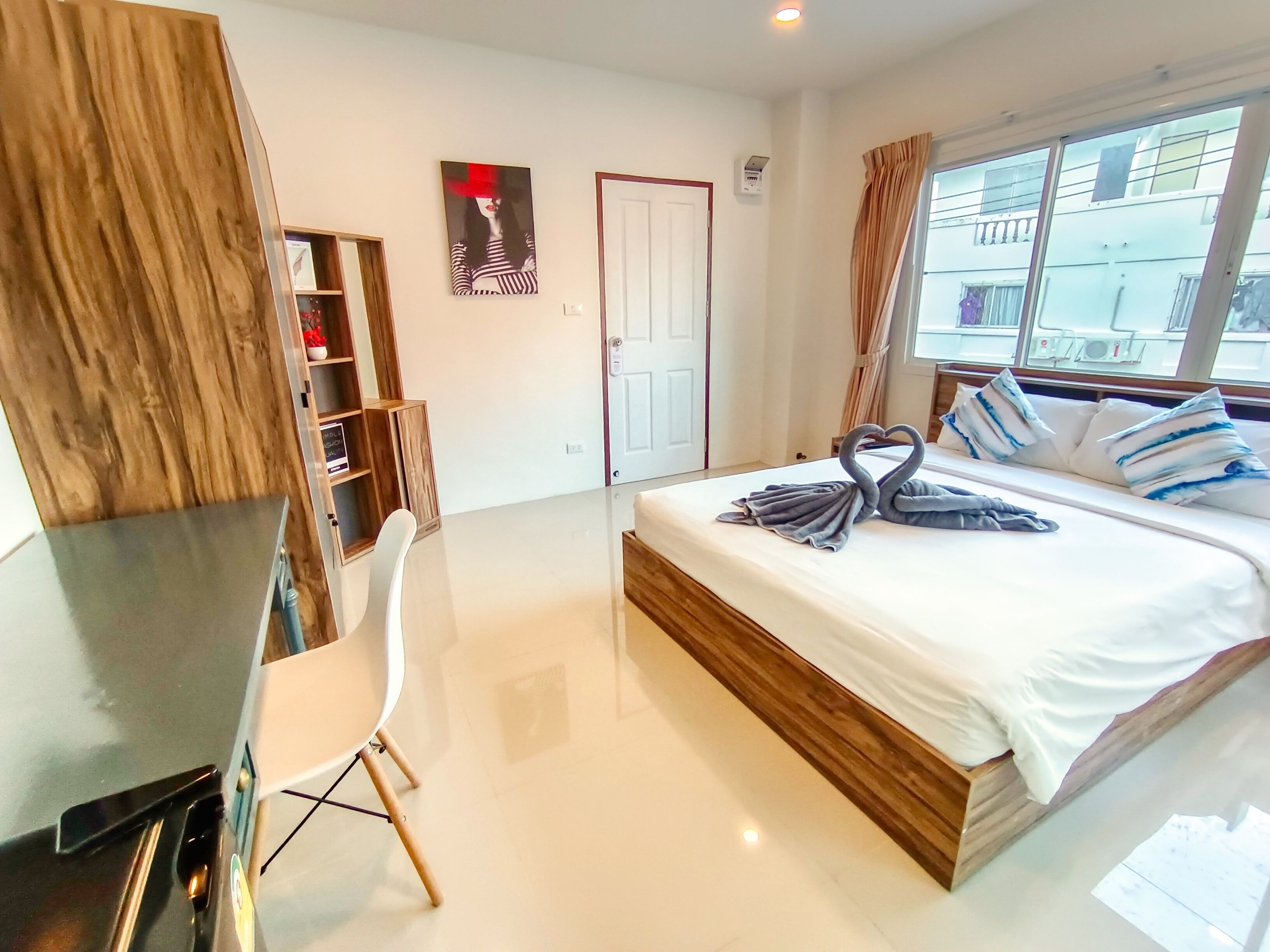 A modern hotel room with a large bed, a desk, and a chair. There is a door, a window with curtains, and a wall-mounted air conditioning unit.