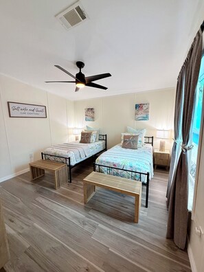 Quarto com duas camas, ventoinha no teto, uma mesinha e janela com cortinas.