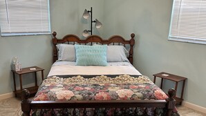 A bedroom with a wooden bed, floral bedspread, two bedside tables, a nightstand, a chandelier, and a window with blinds. - Furnished, lakefront 2/1 house in Ocala Nat'l Forest (Silver Springs)