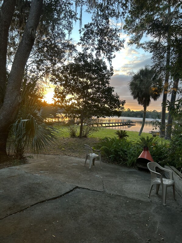 A concrete patio with two plastic chairs, a red planter, and a view of a lake with a dock in the background. - Furnished, lakefront 2/1 house in Ocala Nat'l Forest (Silver Springs)