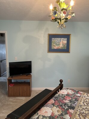 A bedroom with a bed, a TV on a wooden stand, a framed picture on the wall, and a chandelier. - Furnished, lakefront 2/1 house in Ocala Nat'l Forest (Silver Springs)