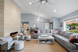 Una sala moderna con un sofá gris, un reposapiés y sillones estampados. Hay un ventilador de techo, una chimenea y vistas al exterior.