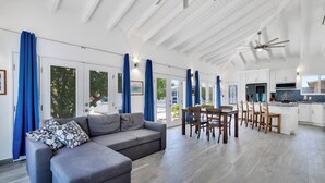 Ein großzügiger Wohnbereich mit grauem Sofa, Esstisch und Stühlen, einer Küchenzeile mit weißen Schränken sowie großen Fenstern mit blauen Vorhängen.