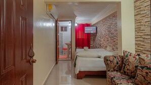 A hotel room with a wooden door, a bed with white bedding, a patterned sofa, a television on the wall, and a bathroom visible through an open door.