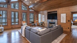 A cozy log cabin interior with a large gray sectional sofa, wooden floors, and large windows offering views of the surrounding forest. - Lake Oconee Lake Front Cabin+Private Dock+Fire Pit+Fireplace+AMAZING VIEWS! (Eatonton)