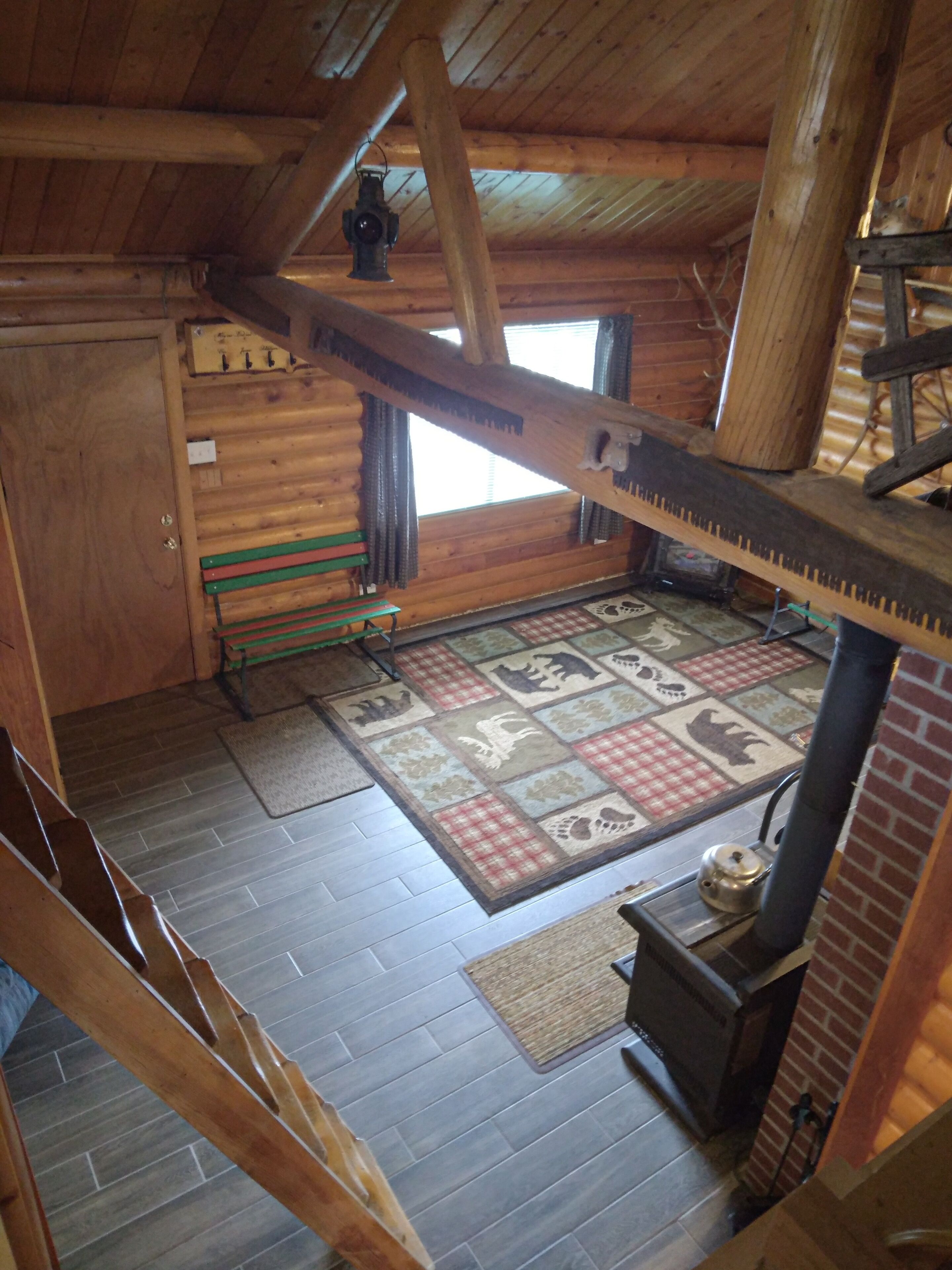 Interior de una cabaña con suelo de madera, alfombra acolchada, banco de madera, estufa y ventana con cortinas.